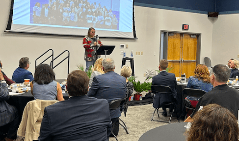 Woman presenting on a stage in front of a crowd of banquet tables.