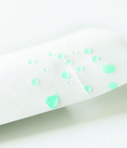 Hydroblox, JBM"s water resistant paper packaging. A white piece of paper is shown with beaded droplets of aquamarine liquid.