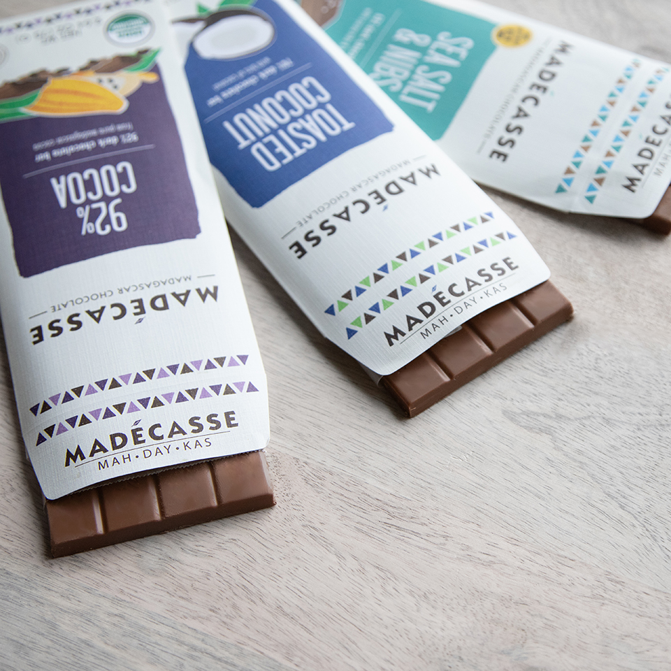 Three Madecasse chocolate bars laid side by side, featuring white packaging with the brand name in bold. There are three different flavors featured: "Sea Salt and Nibs" in a teal color, "Toasted Coconut" in a royal blue, and "92% Cocoa" in a deep purple.