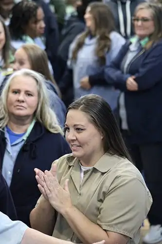 A woman in a prison uniform is smiling and gesturing with her hands, with other incarcerated women in the background.