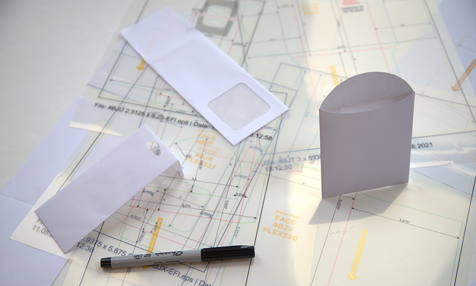 Three paper bags and envelopes in the package design phase with drawings of dimensions in the background, showcasing how custom bags are created at JBM.