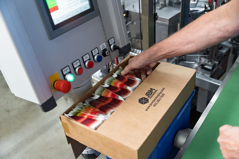 Hand reaching down into a JBM cardboard box filled with stacked seed packets, resting on a machine with a screen and colorful buttons.
