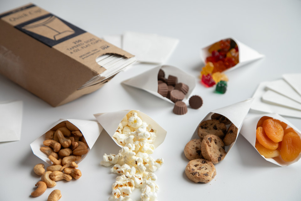 Small, foldable paper cups hold nuts, cookies, popcorn, and other snacks. A cardboard package holding hundreds of flat paper cups is nearby.