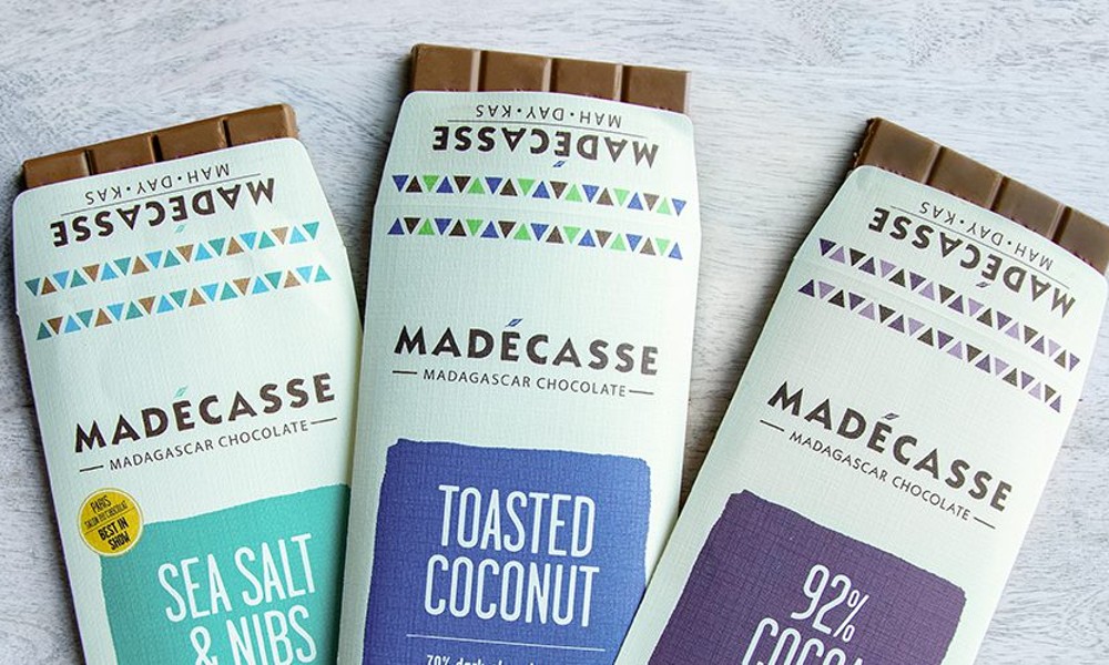 Three Madecasse chocolate bars laid side by side, featuring white packaging with the brand name in bold. There are three different flavors featured: "Sea Salt and Nibs" in a teal color, "Toasted Coconut" in a royal blue, and "92% Cocoa" in a deep purple.
