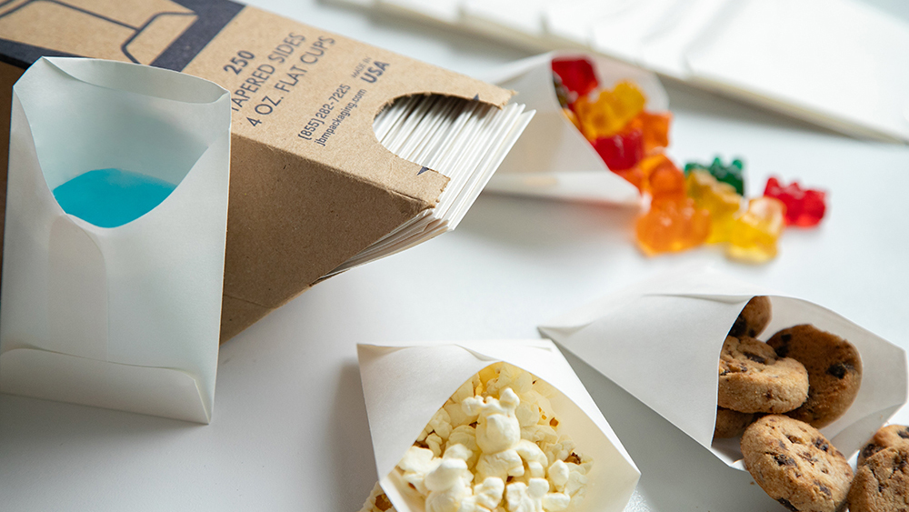 White, flat paper cups holding a variety of food and beverage items like popcorn, blue beverage, gummy bears, and miniature chocolate chip cookies.